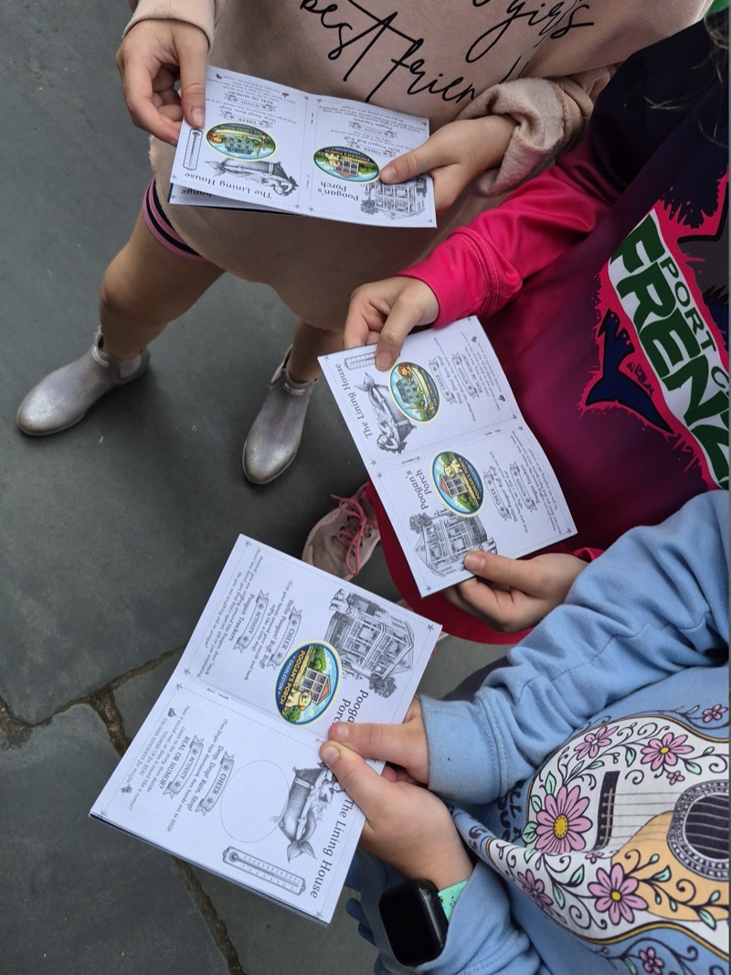 Three children on a Charleston sidewalk holding open their Junior JWalker passports, each page showing an illustrated stop — Pogue's Porch and The Lining House — with a color sticker earned beside it