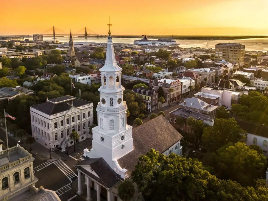 Aerial view of Charleston at sunset: a white church steeple rising above the historic district, with the harbor, the Ravenel Bridge and a cruise ship beyond